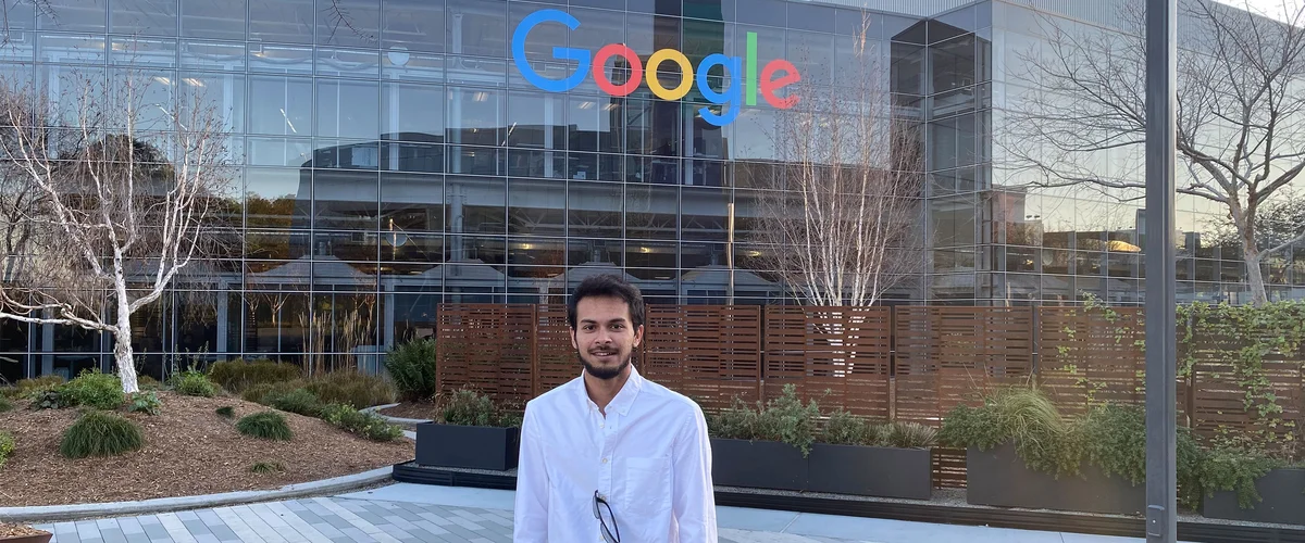 Akash stands in front of a Google building with a multicolor Google logo across the top. In the background is landscaping and a fence.