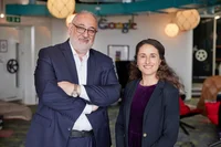 A man in a blue suit and glasses and a woman with dark curly hair and a black shirt stand beside one another. Behind them is an office with large overhead lights and the Google logo.