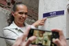 A woman smiles while pointing to a board fixed to a residence with a digital address