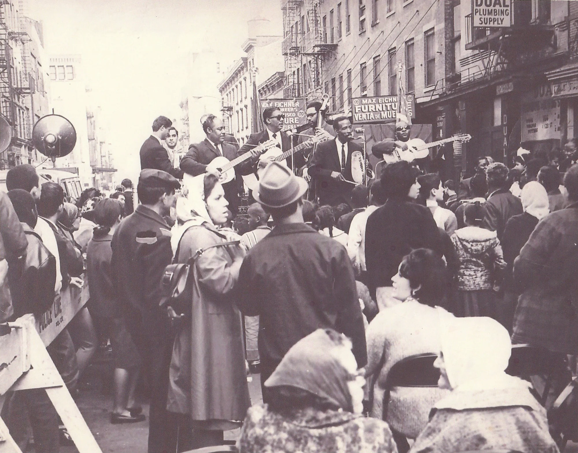 Black and white photograph of a crowd watching Alfonso Panama and his band perform in the middle of the street in NYC in 1957. The crowd is a mix of men and women, watching 7 men play cuatro, the güira, and the double bass while singing.