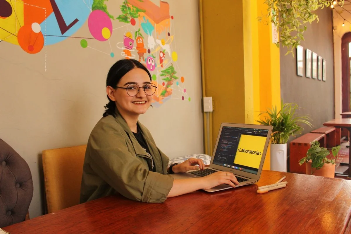 Mulher de óculos estuda no computador e sorri para a foto. Logo do Laboratória em destaque no monitor.