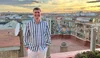 A man with a blue-and-white striped shirt and white trousers stands on a roof deck at sunset with a cityscape in the background