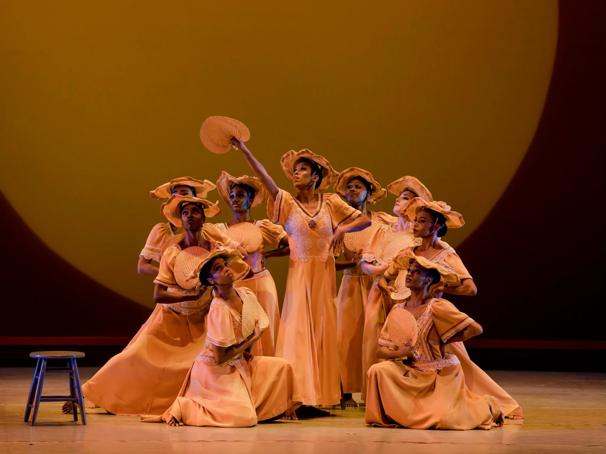 Color photograph of nine women standing on a stage, surrounded by three dark-wooded stools. The women wear long, yellow-colored gowns, wear straw sun hats, and have straw fans in their hands. One woman extends her arms, fan in hand, to what they all have their gaze set to.