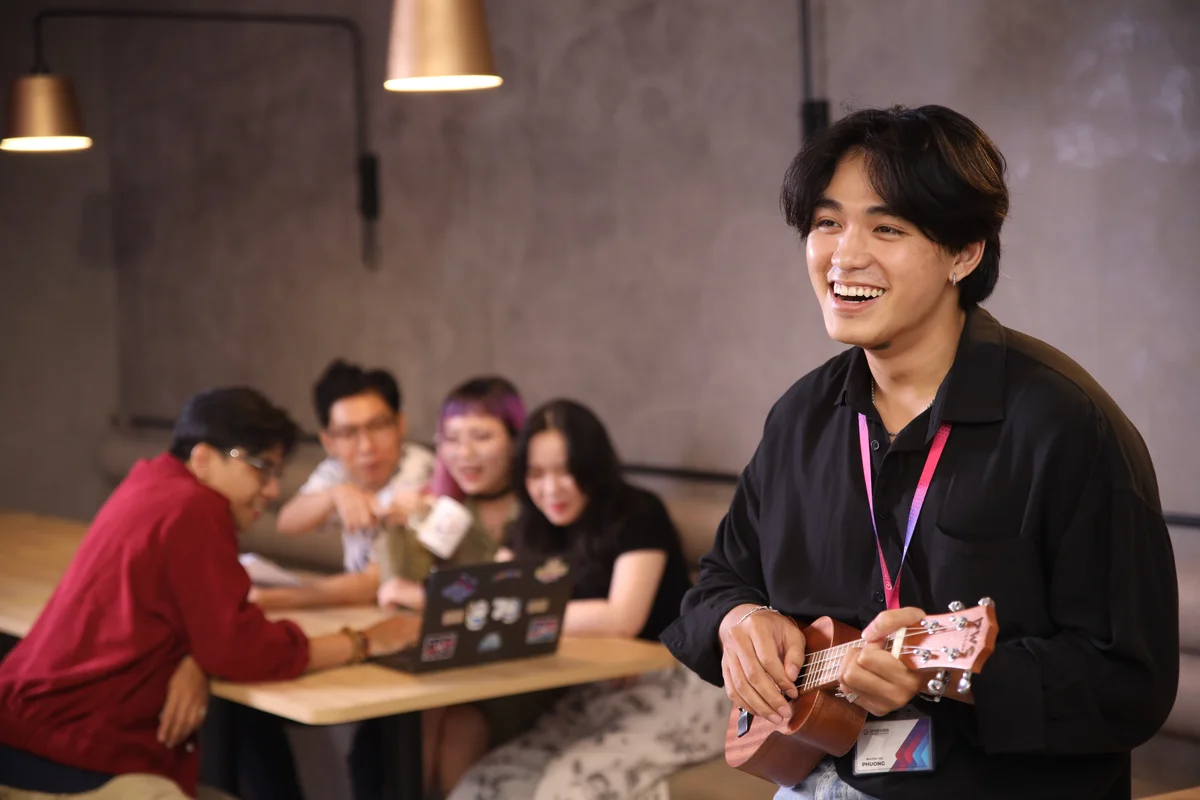 A close-up of a man smiling and playing the ukulele. Behind him are four people gathered around a laptop talking.