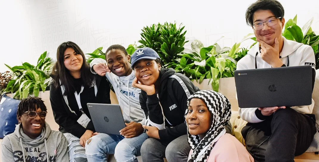 A group of people sitting on a bench and smiling at the camera, two people with laptops.