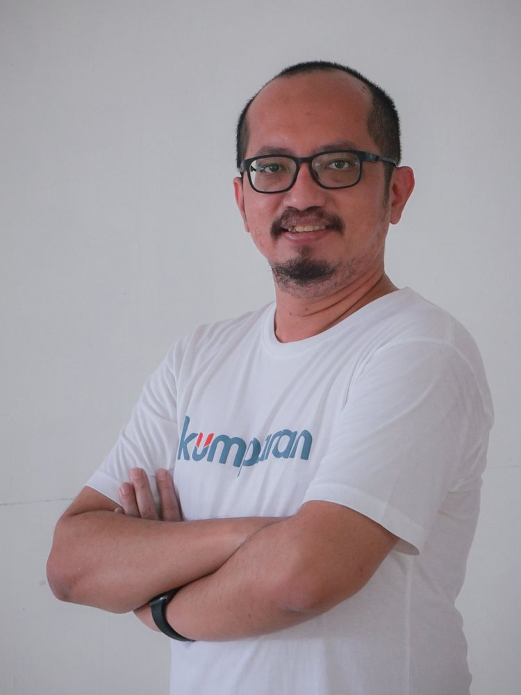 This is a photograph of the Chief of Corporate Strategy at kumparan, called Andrias Ekoyuono. He is standing up and looking directly at the camera wearing a white T-shirt with the kumparan logo across the chest in blue with orange highlights.