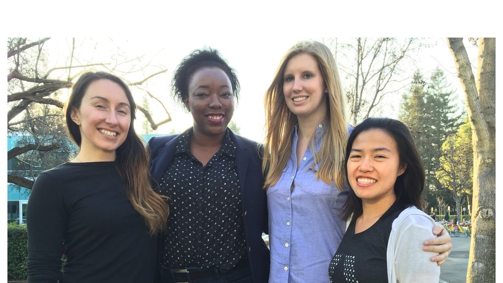 Photograph of four women standing with their arms around each other, looking into the camera and smiling.