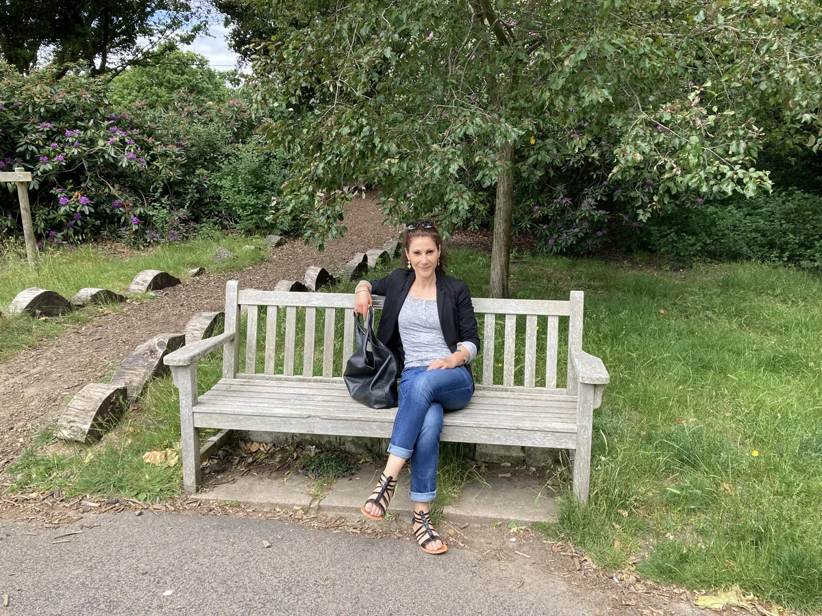 A woman wearing jeans and a black jacket sitting on a bench at a park with trees behind her.