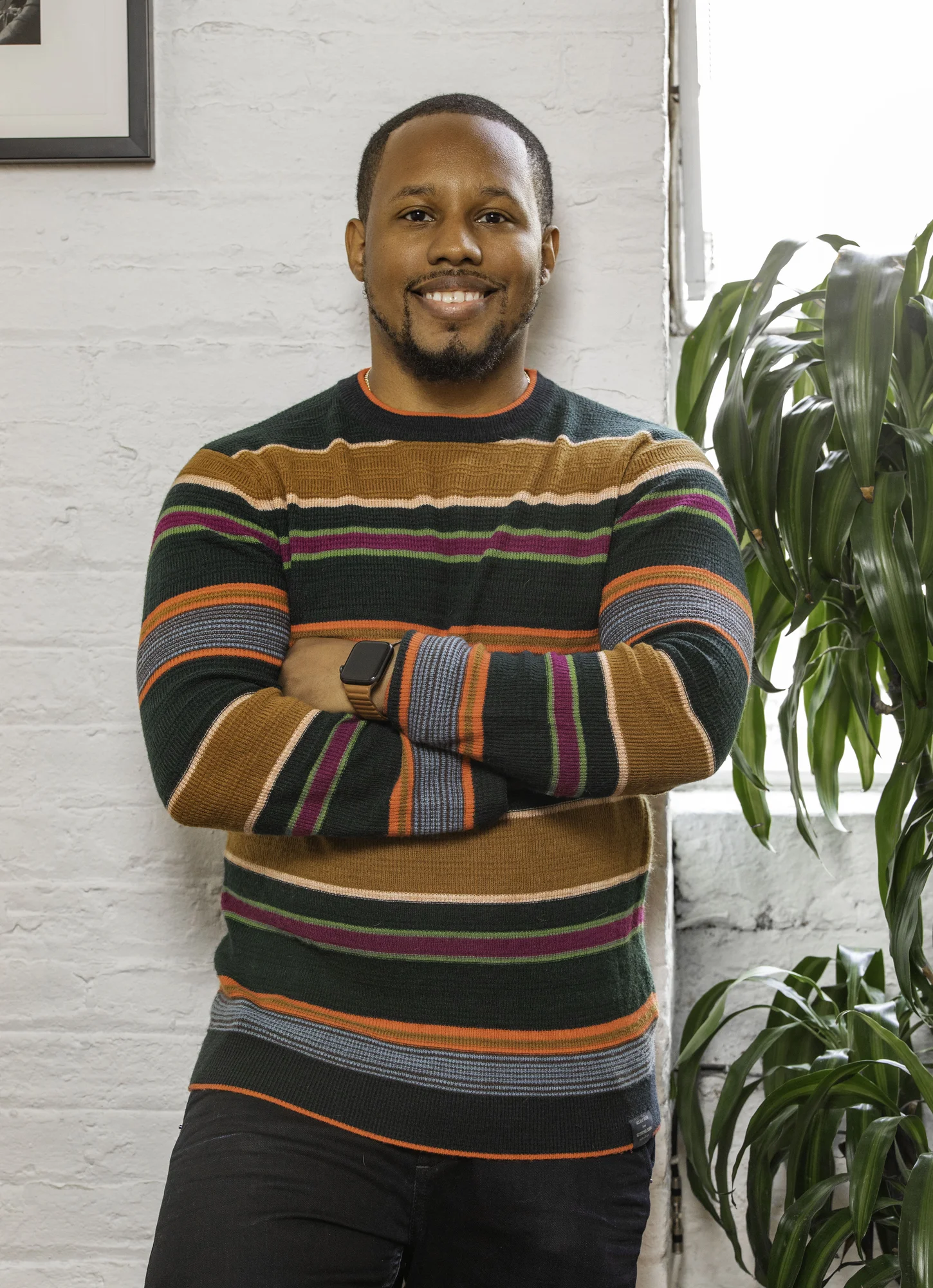 Anthony Edwards Jr stands with his arms crossed in a vivid sweater with orange, green, teal, salmon and cream stripes. He smiles at the camera and leans against a wall behind him.
