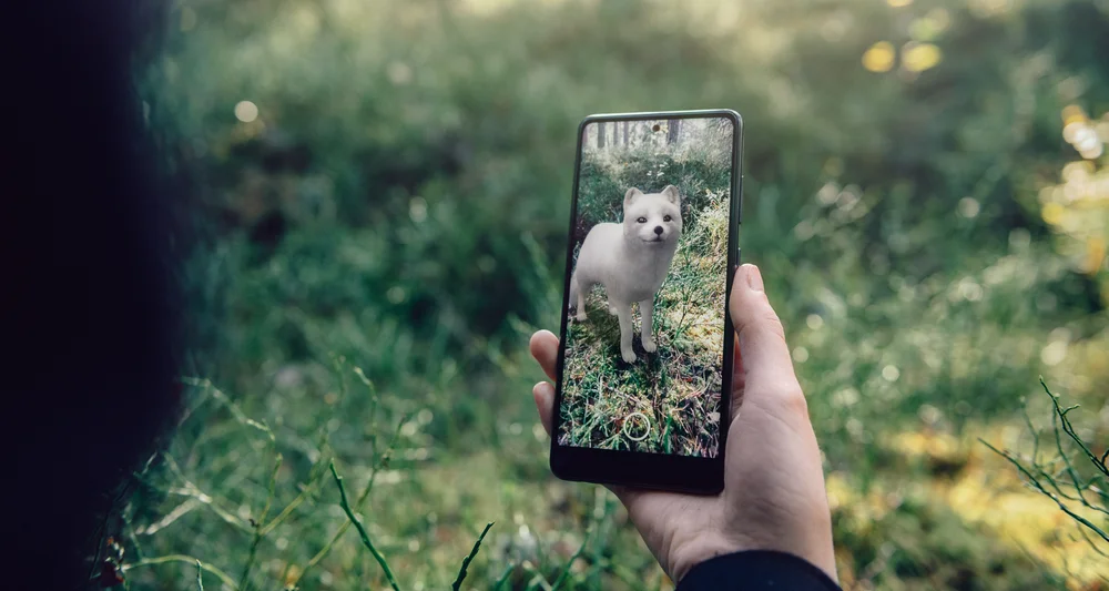 A woman is holding a phone with a augmented reality image of a white arctic fox superimposed on the green landscape in front of her
