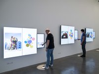 Photograph of an installation composed of nine touch screens, organized in three groups (two large screens and a smaller one on the right). Two men are touching two of the small screens.
