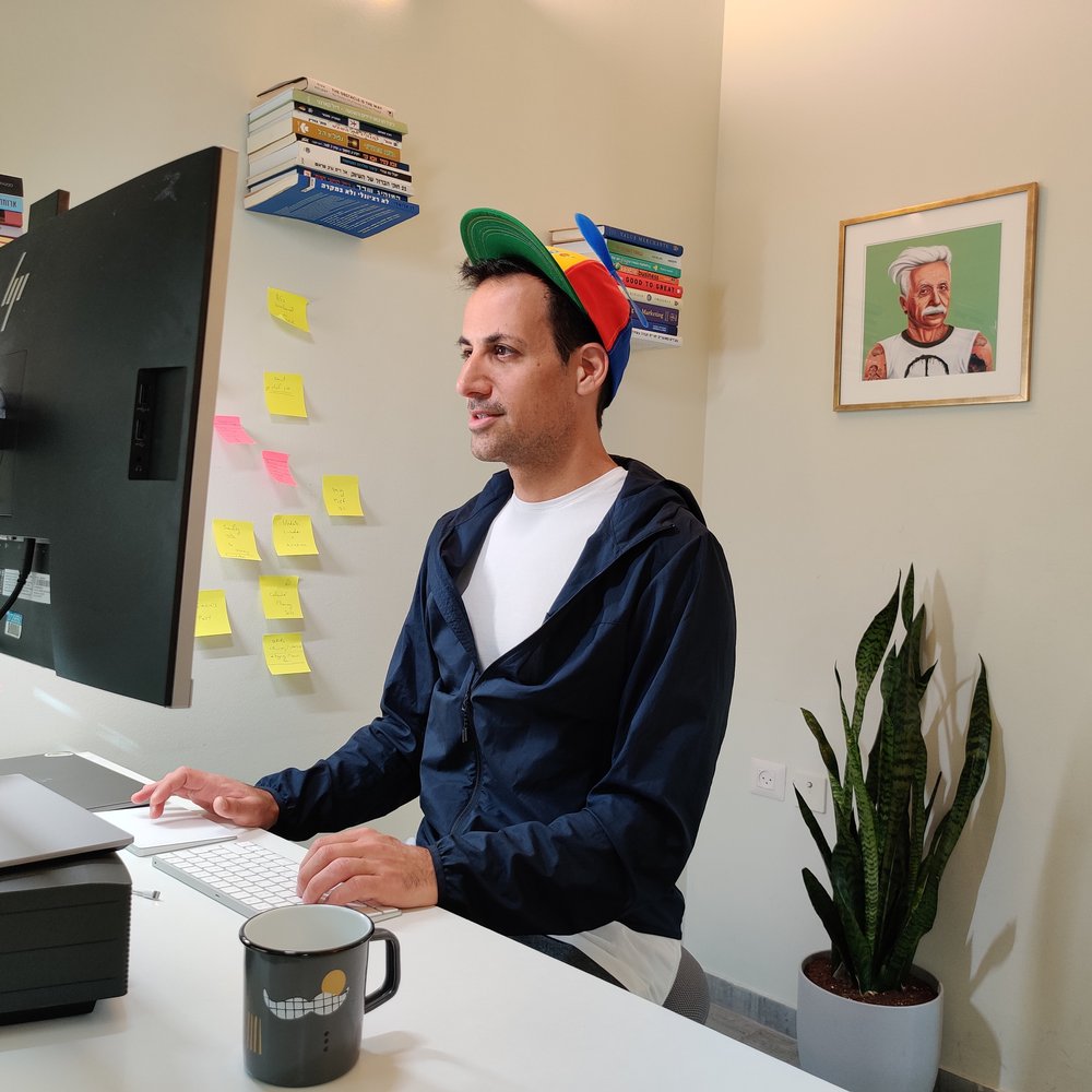 Asaf wearing a Noogler hat while sitting in front of a computer. Around him are books, sticky notes on a wall, a mug, a plant and a painting.