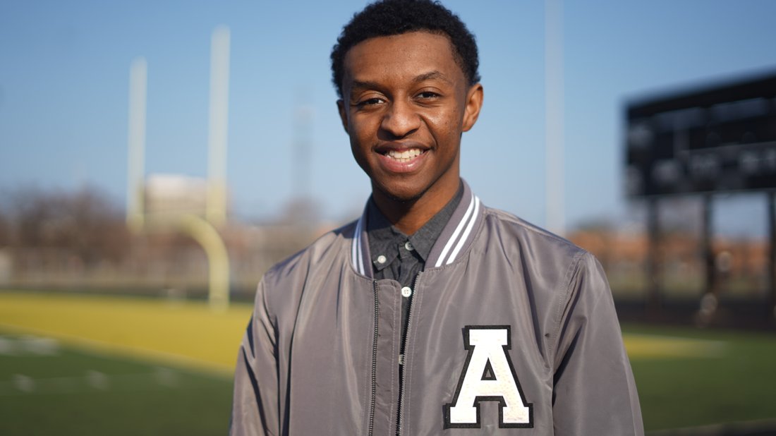 Ashton Keys smiles at the camera wearing a gray polo and gray letterman jacket with a large, white, “A” on it. Behind him, a football field is visible.