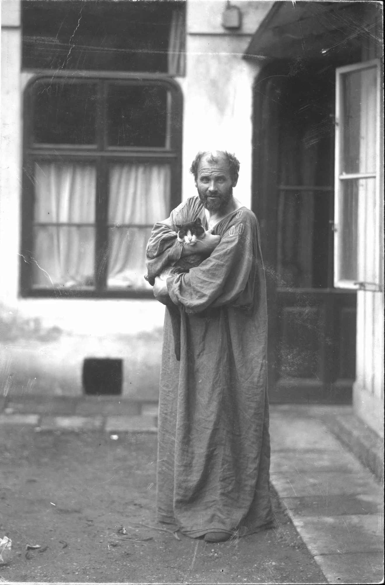 Black-and-white-photograph of Gustav Klimt standing in front of his studio, holding a cat on his arm