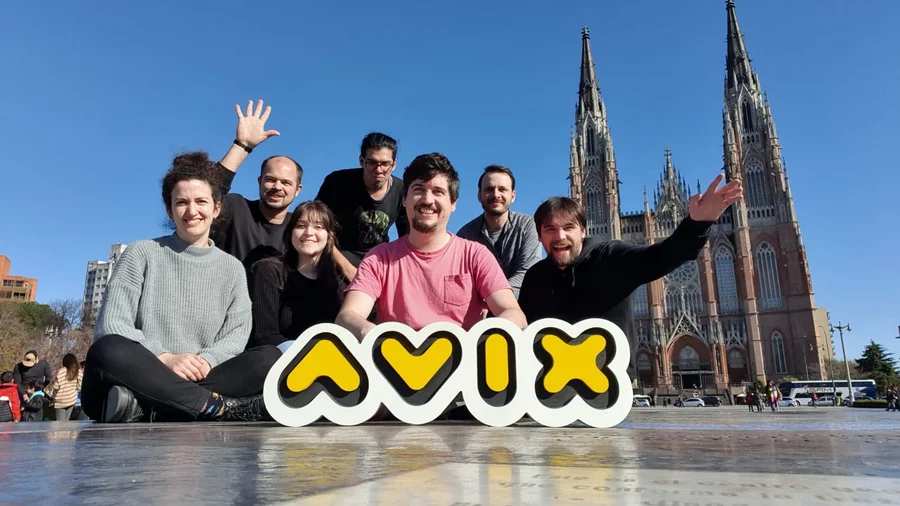 Seven people sit on the ground in front of a cathedral. Some are smiling and waving, and the person in front is holding a large sign that says “Avix.”