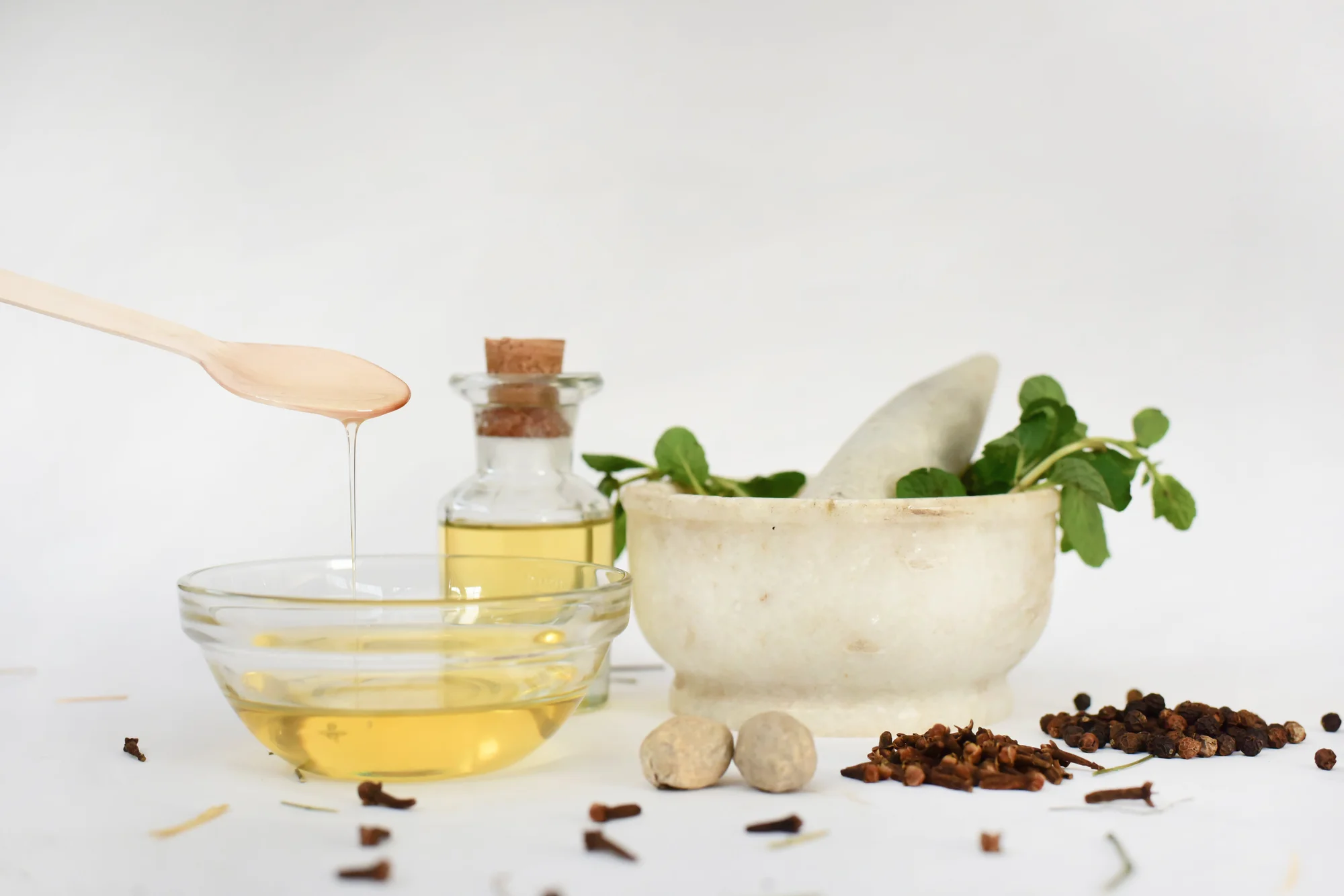 Color photograph of a glass bowl of honey, mortar and pestle with green leaves, a vial of yellow clear liquid, and various loose spices against a white background.