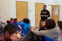 A Googler stands in the front of a room holding a tablet, smiling at a group of young students as they use