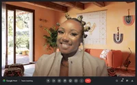 A woman sitting in a virtual meeting hosted on Google Meet. The virtual background is set in a living room with color tones of orange and tan. In the background there is  symbolism from the Sankofa bird, vibrant textiles, jazz instruments and more.