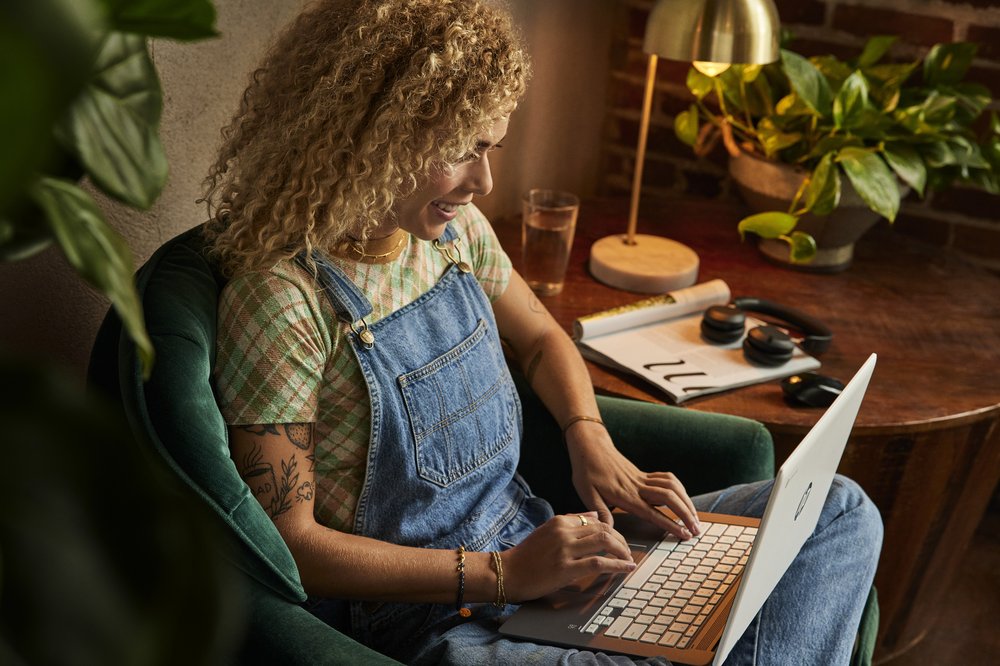 A woman wearing overalls sits in a green chair with a Chromebook on her lap.