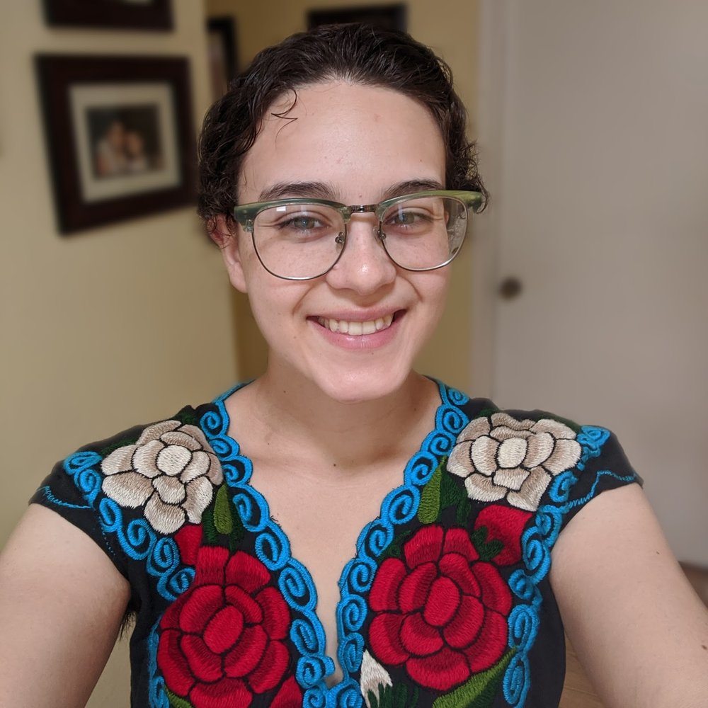 Image shows a young woman taking a selfie; she's looking into the camera and smiling. She has dark hark and glasses and is wearing a shirt with flowers on it.