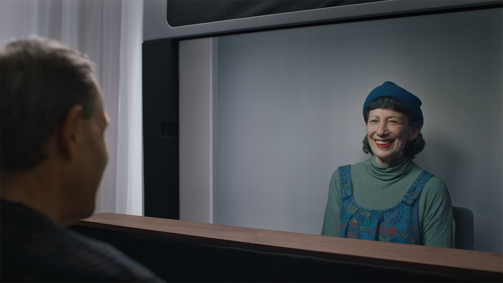 A woman wearing a green shirt, blue overalls and a blue hat smiles on screen as a man, seen in profile, watches.