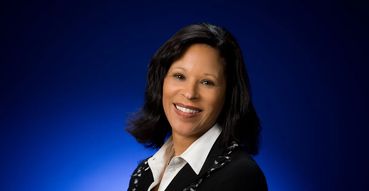 Image shows a headshot of a woman against a blue background; she is looking at the camera and smiling.