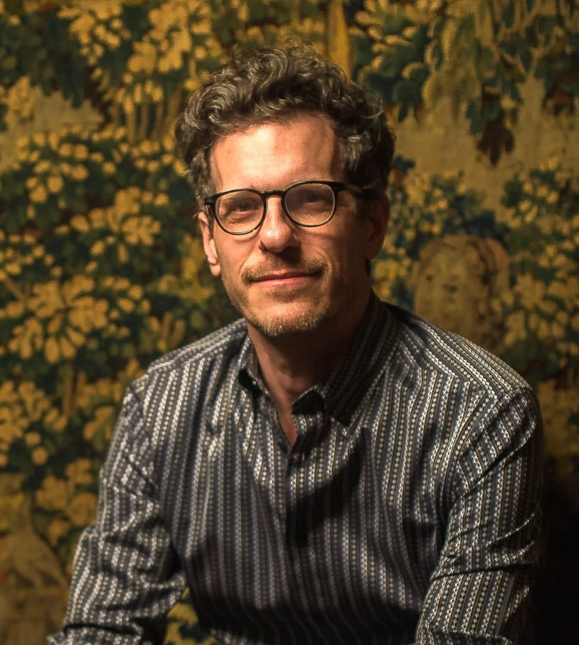 Photograph of  Brian Selznick smiling directly at camera in a striped button up shirt and black framed eyeglasses. Brian has white skin and curly, dark, short hair.