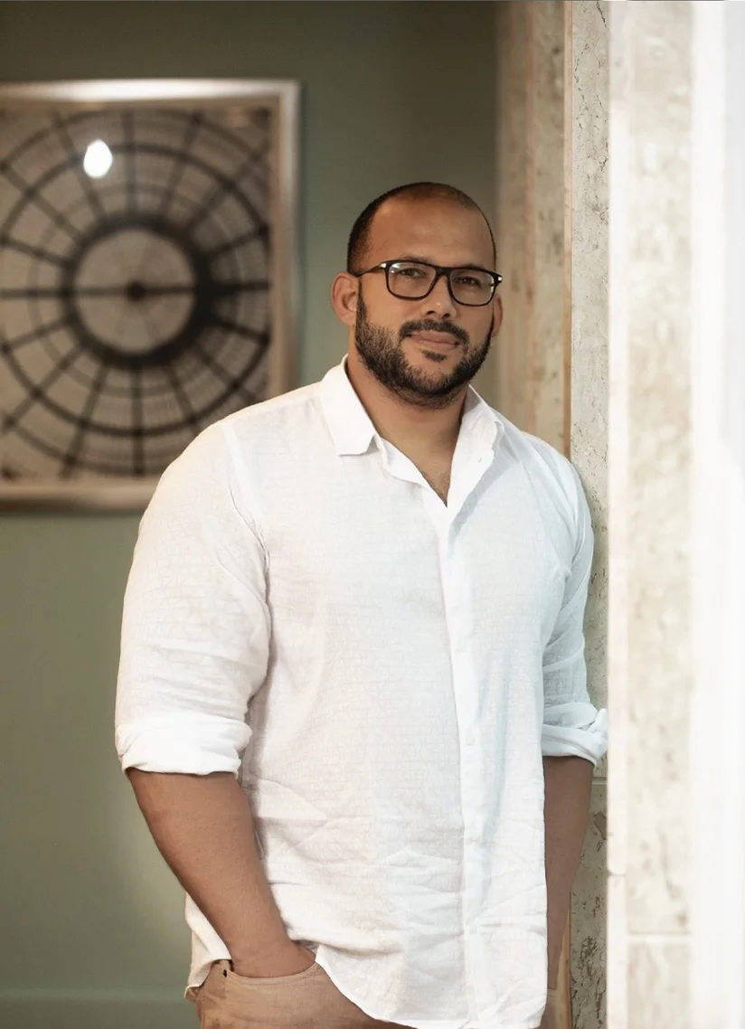 A man with facial hair and glasses looks at the camera, one hand in his pocket. He's wearing a white shirt and standing near a window with white curtains, in front of a green wall.