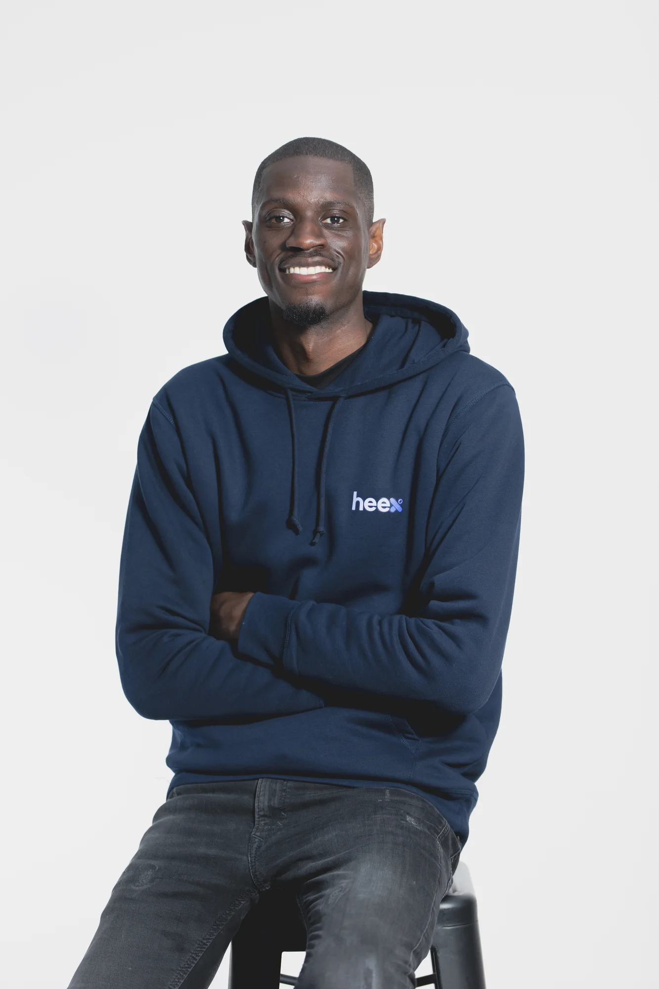 A man wearing a navy blue sweatshirt and jeans sits on a stool, looking at the camera and his arms folded across his chest.