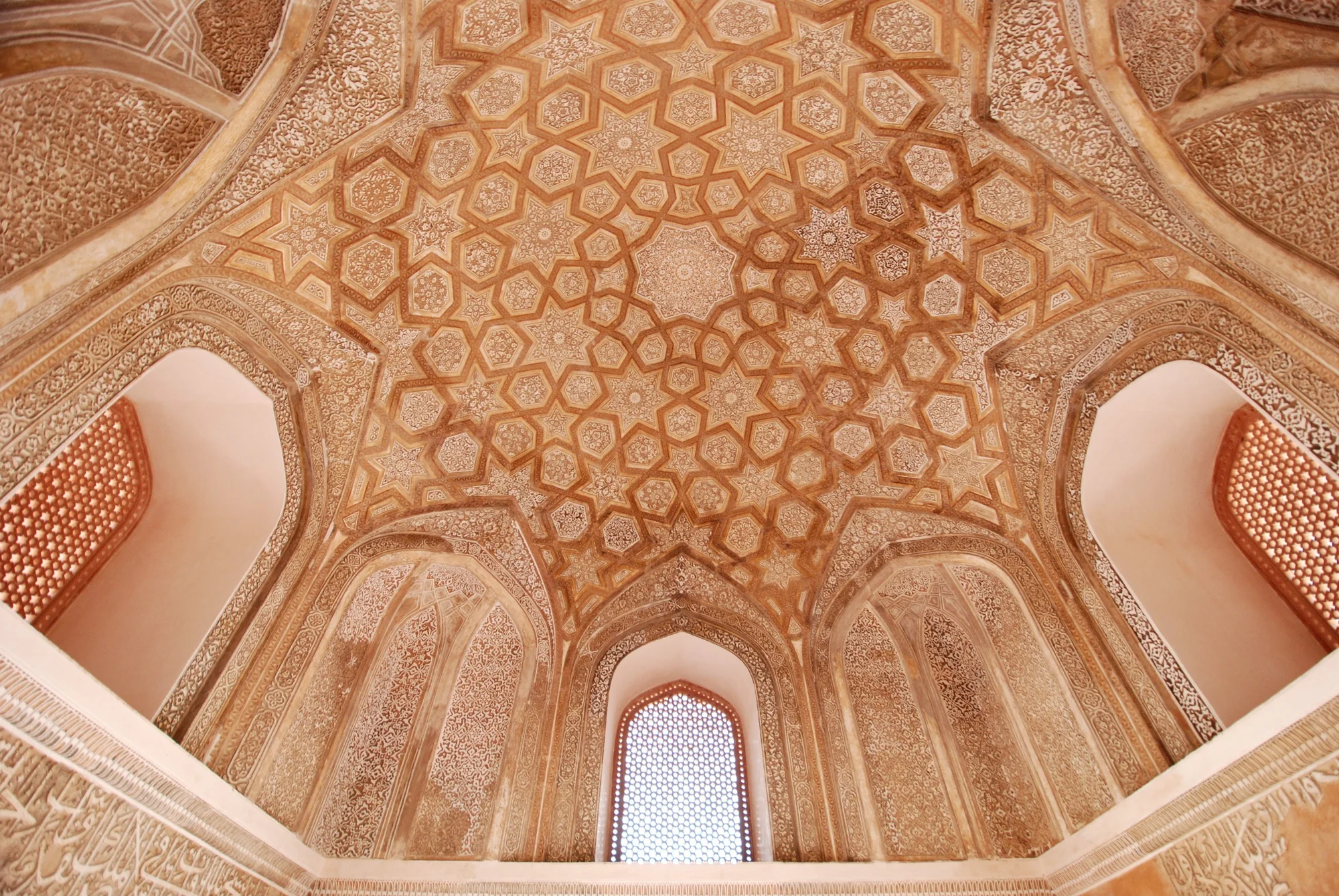 A view of a dome ceiling with elaborate red and earth tone patterns. There are seven niches shown, three of which are windows with geometric-patterned window coverings.