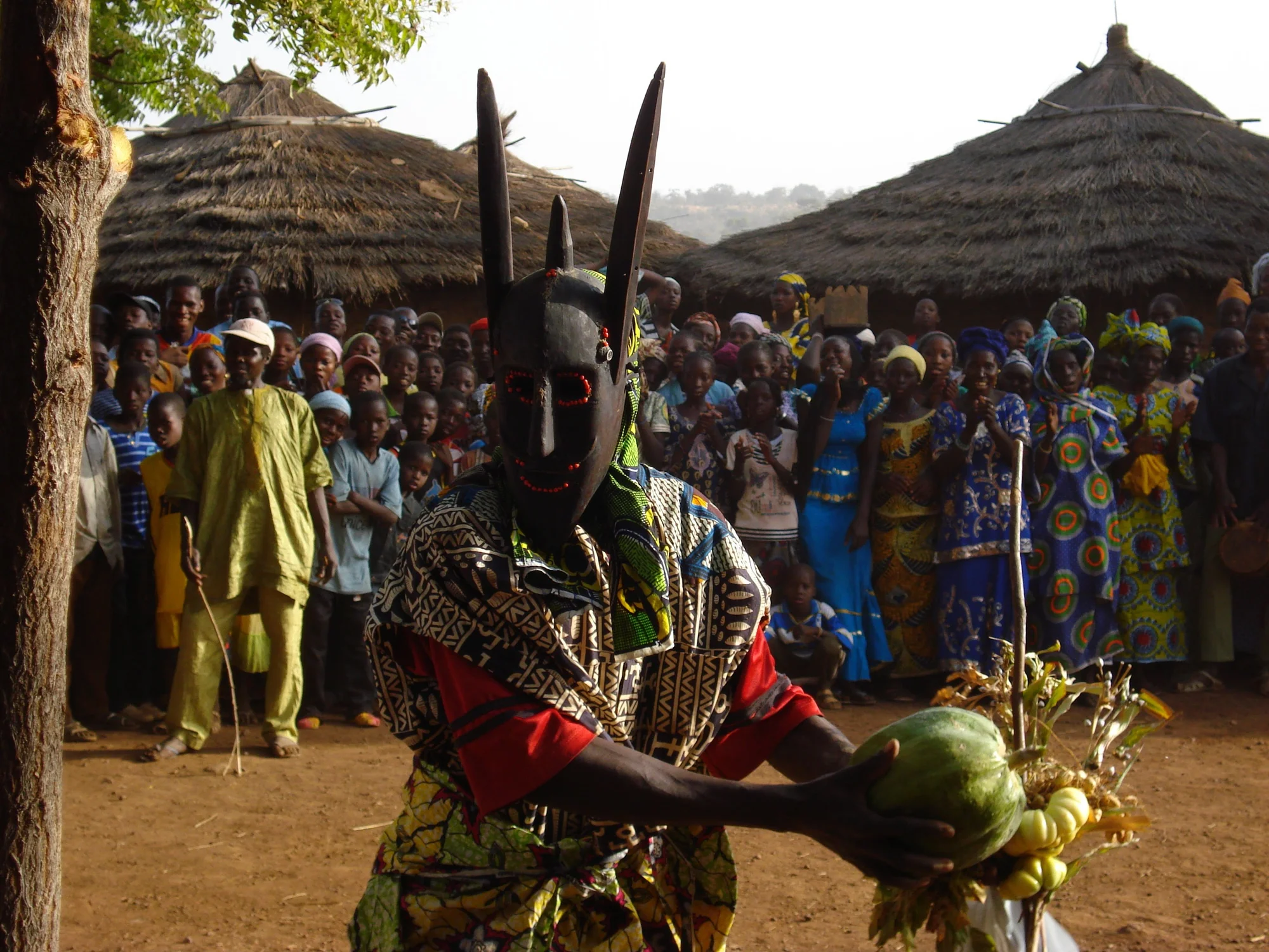 A masked man in colorful clothing, holding a variety of pumpkins to his side, in the middle of a performance watched by an audience behind him.