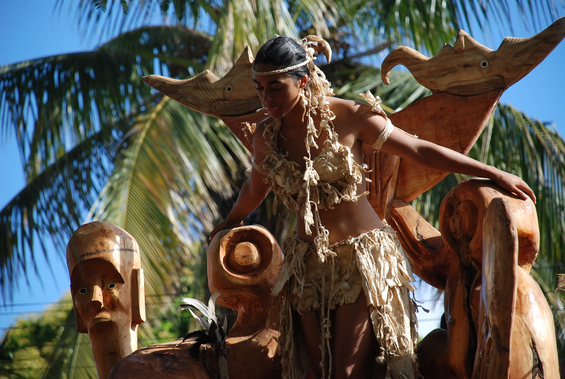 A young person takes part in a festival on Rapa Nui. They are outside with palm trees and a blue sky behind them.