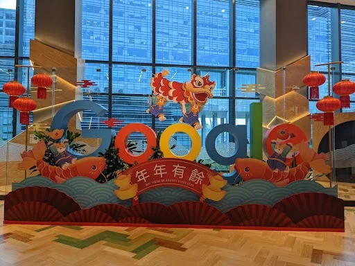 A Google sign in a lobby is decorated for Lunar New Year with pink, red and white flowers. On the right is a tiger standing with a pile of gold pieces.