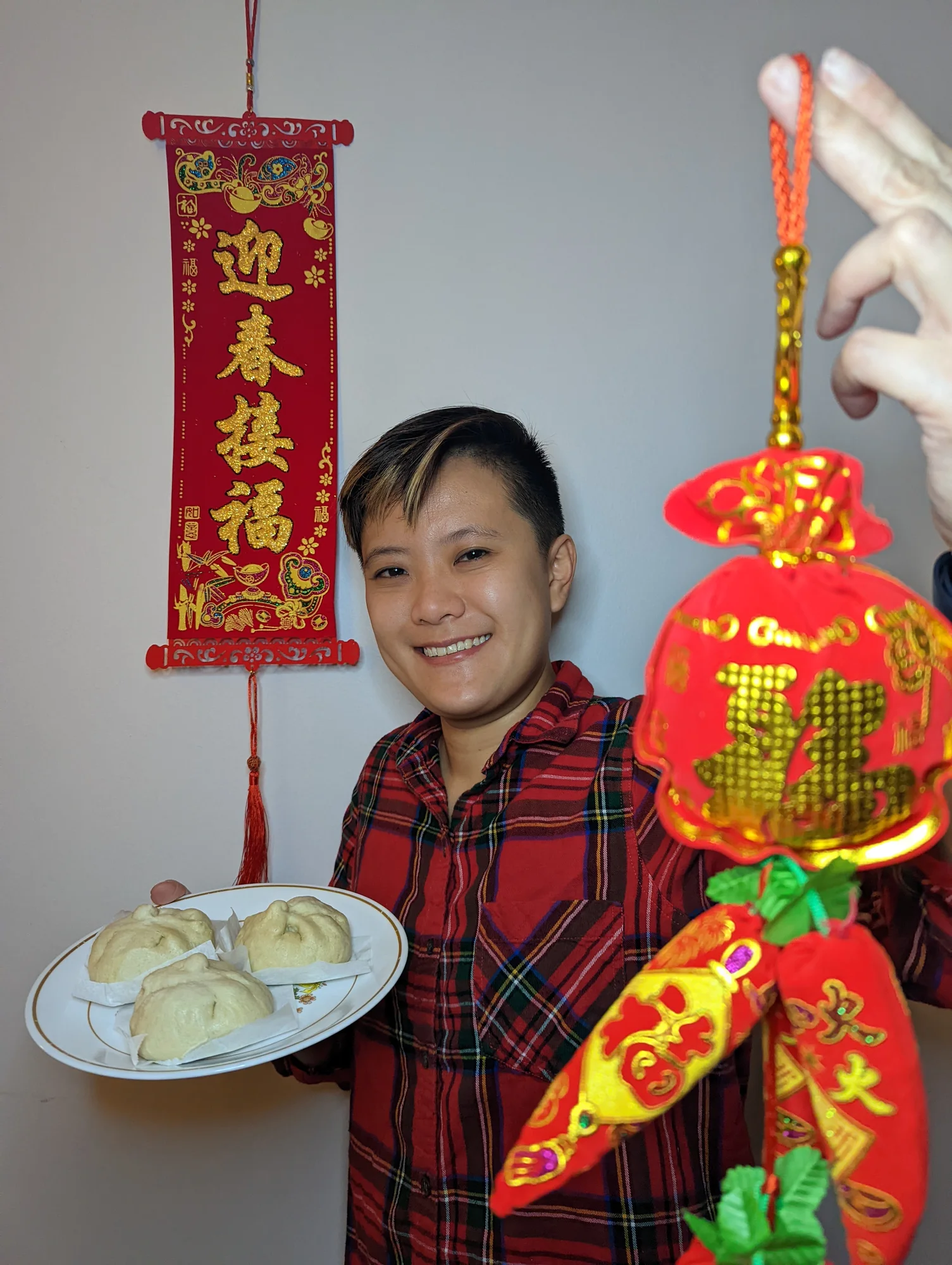 A photography of a man smiling into the camera holding a plate of dumplings and surrounded by red hanging decorations.