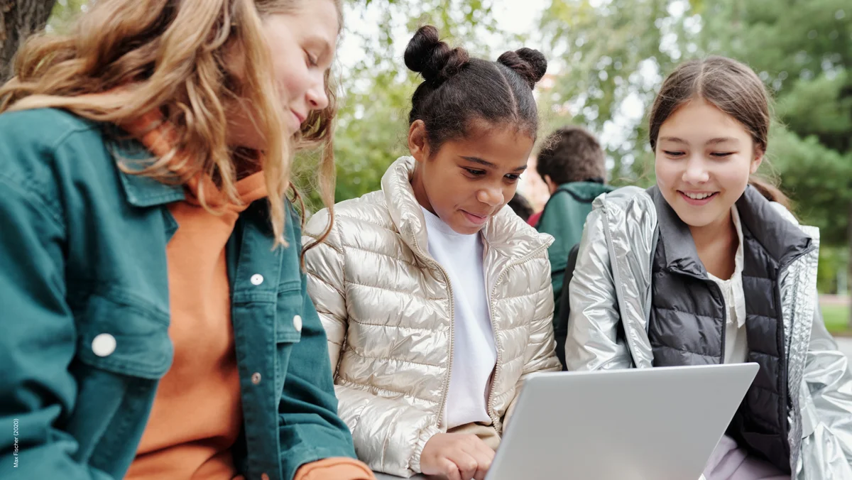 Three young girls are sitting down side by side, with a laptop on the lap of the middle girl