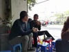 Sundar Pichai in conversation with “Flappy Bird” creator Dong Nguyen. They are sitting on low stools in a street-side cafe in Hanoi, with motorcycles, pedestrians, trees and shops in the background. A smartphone is propped on a stool in front of them.