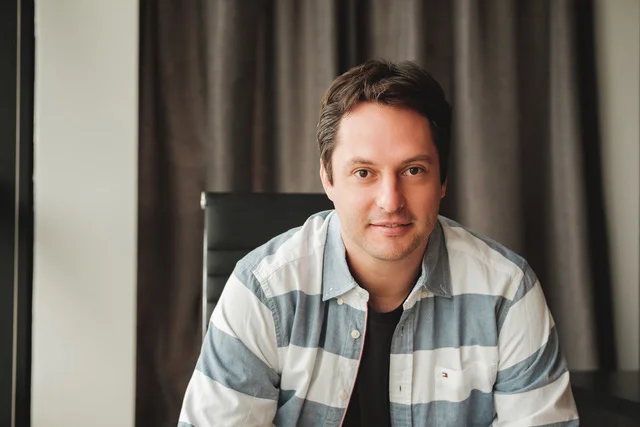 A man in a striped button-down shirt sits on a chair and smiles.