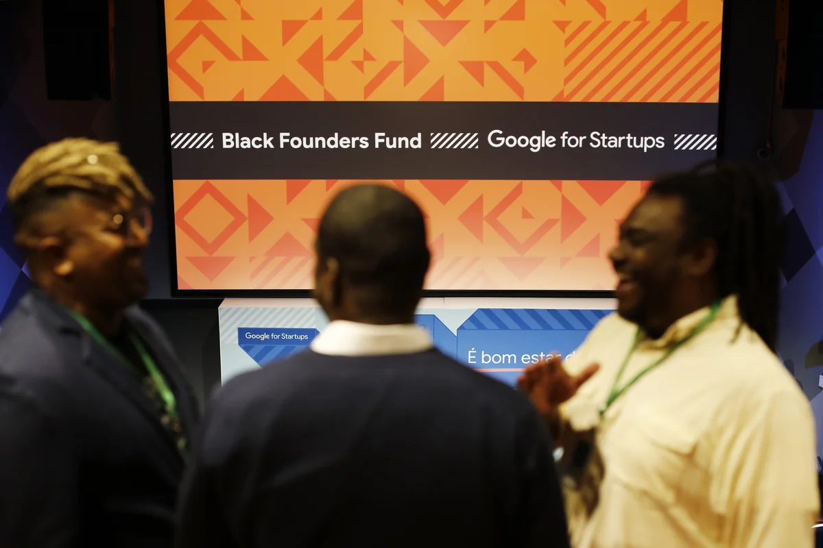 Três homens negros conversam em frente a um monitor grande apresentando o título Google for Startups