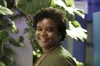 A Black woman with curly short hair and wearing a green top smiles at the camera. In the background is a blue wall and several large indoor plants.