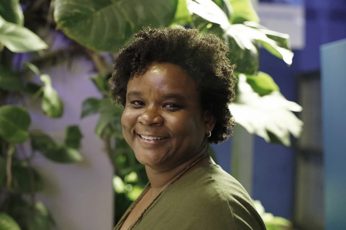 A Black woman with curly short hair and wearing a green top smiles at the camera. In the background is a blue wall and several large indoor plants.