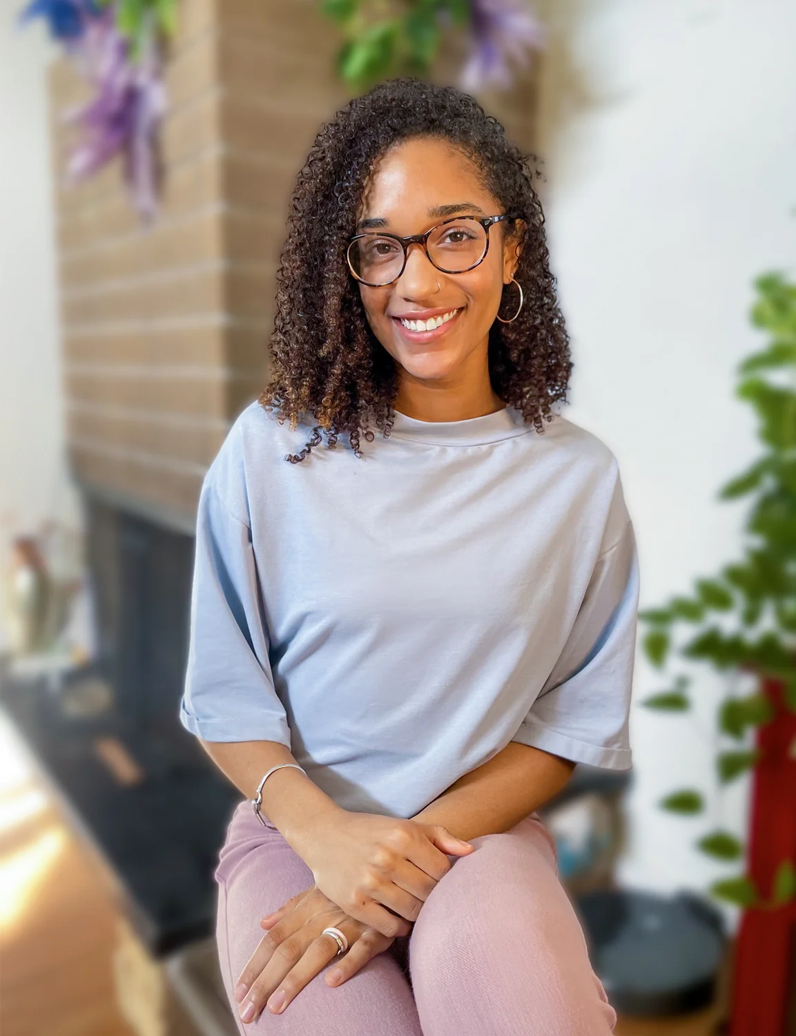 Candice Speicher smiles at the camera while seated in a gray blouse and mauve pants. She wears glasses and hoop earrings.