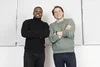 A Black man wearing a black turtleneck and black pants stands beside a white man wearing a sage green sweater and blue jeans. Both mean have their arms crossed and are facing the camera.