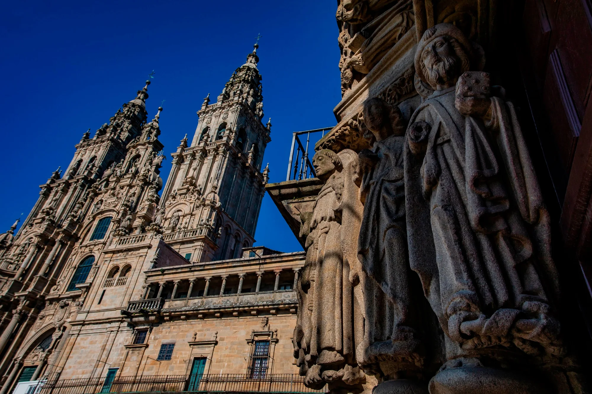 A photograph of the Cathedral of Santiago de Compostela in which can be seen in the foreground, two stone statues, and in the background, the facade of the Obradoiro of the Cathedral.