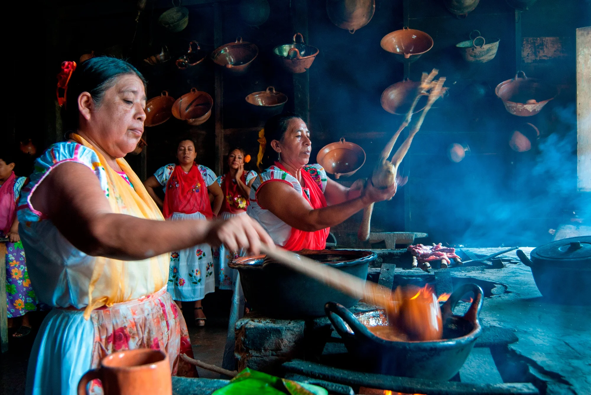 Two women cook together with three women observing behind them. The woman in the foreground uses a ladle to stir soup. The other woman is preparing meat above a grill. Pottery is hanging from the wall behind them.