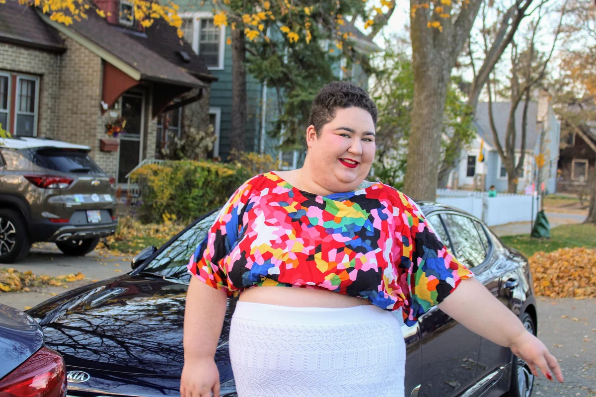 A woman with short brown hair wearing a colorful midriff-baring top and white patterned skirt poses in front of a black sedan in a residential neighborhood.