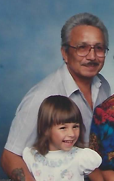 An older man with brown skin and white hair and a mustache in a pale blue button down short sleeve shirt (Cheryl’s Misho) is holding a younger girl (Cheryl) with brown hair and white skin, in a white short, puffy sleeved shirt. They are both smiling at the camera (Misho with a closed mouth smile, Cheryl with a tooth-smile).