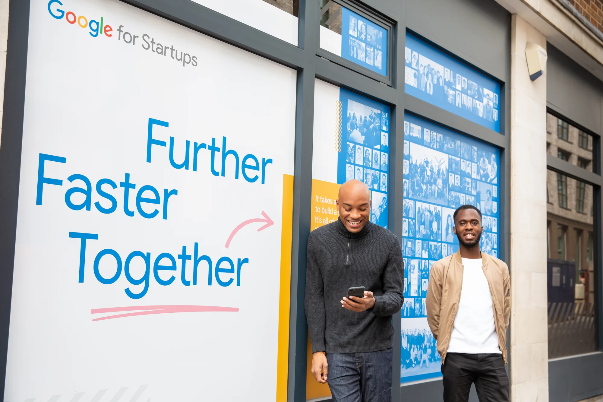 Two men stand against a wall that says "Google for Startups, Further, Faster, Together." One of them looks at his smartphone, smiling, while the other looks into the camera, smiling.