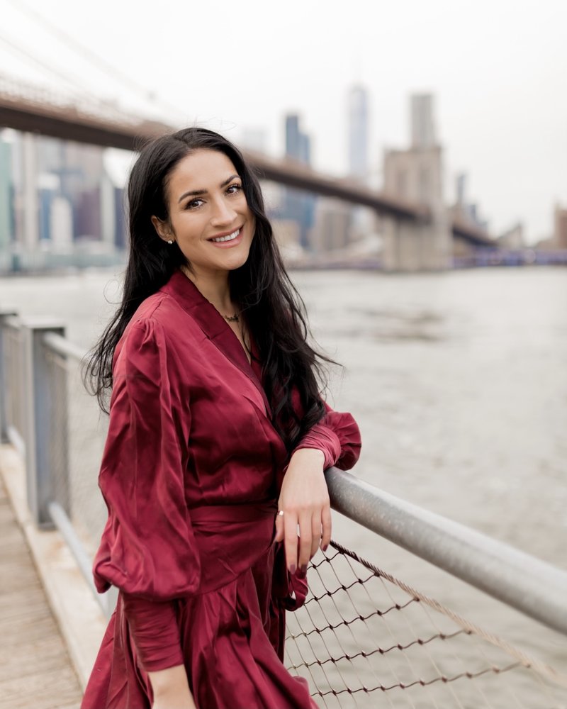 Christina leans against a railing overlooking the water and the New York City skyline. She is smiling with long brown hair, wearing a flowy, long-sleeved red dress.