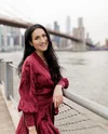 Christina leans against a railing overlooking the water and the New York City skyline. She is smiling with long brown hair, wearing a flowy, long-sleeved red dress.