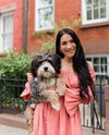 Holding her dog, Koa, and smiling, Christina stands on the sidewalk with a wrought-iron fence and brick building behind her. She has long brown hair down and wears an off-the-shoulder, long-sleeved peach-colored dress.